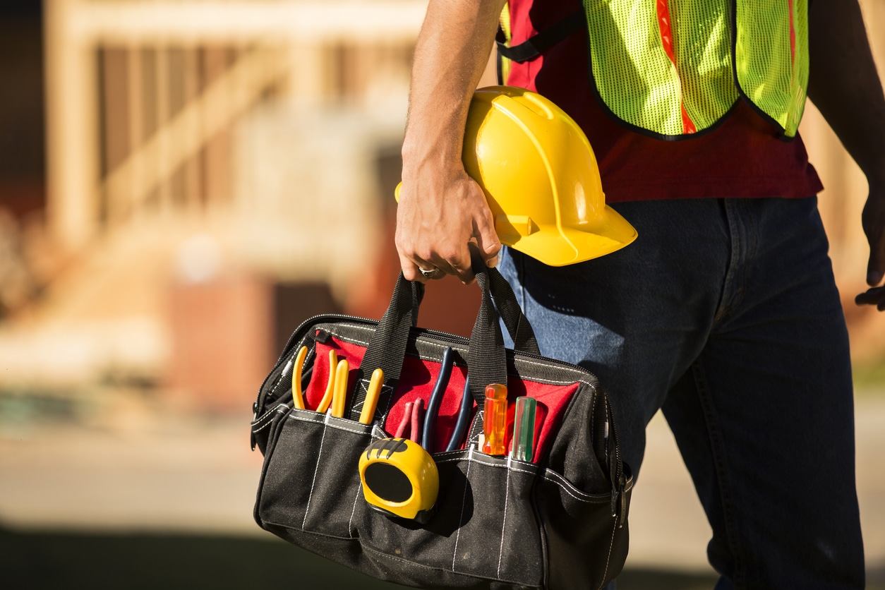 A construction worker busy working at a job site. He holds a tool box full of tools and a hard hat. Framed house, building in background. He is wearing a safety vest.